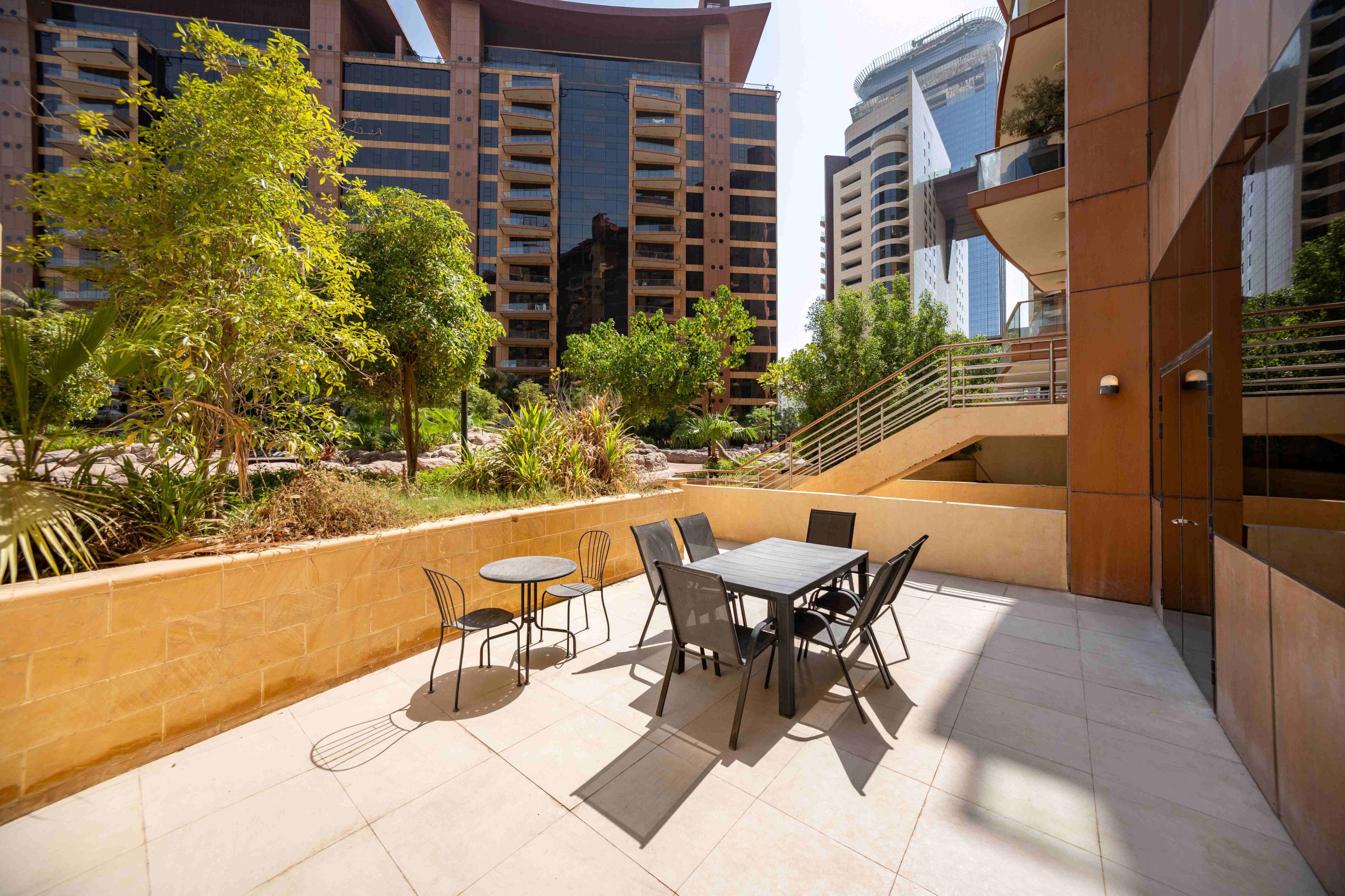 An outdoor patio area with a table and chairs, surrounded by greenery and modern buildings.