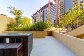 A modern outdoor area with a glass-top table and wicker furniture, surrounded by greenery and a stone wall.