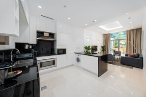 A modern kitchen with a built-in oven, microwave, and washing machine, a dining area with a sofa and table, and a large window with curtains.