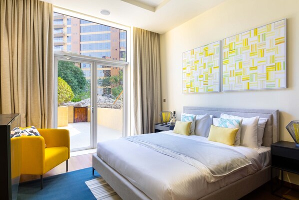 A modern hotel room with a large bed, a yellow armchair, and a view of the city through the window.