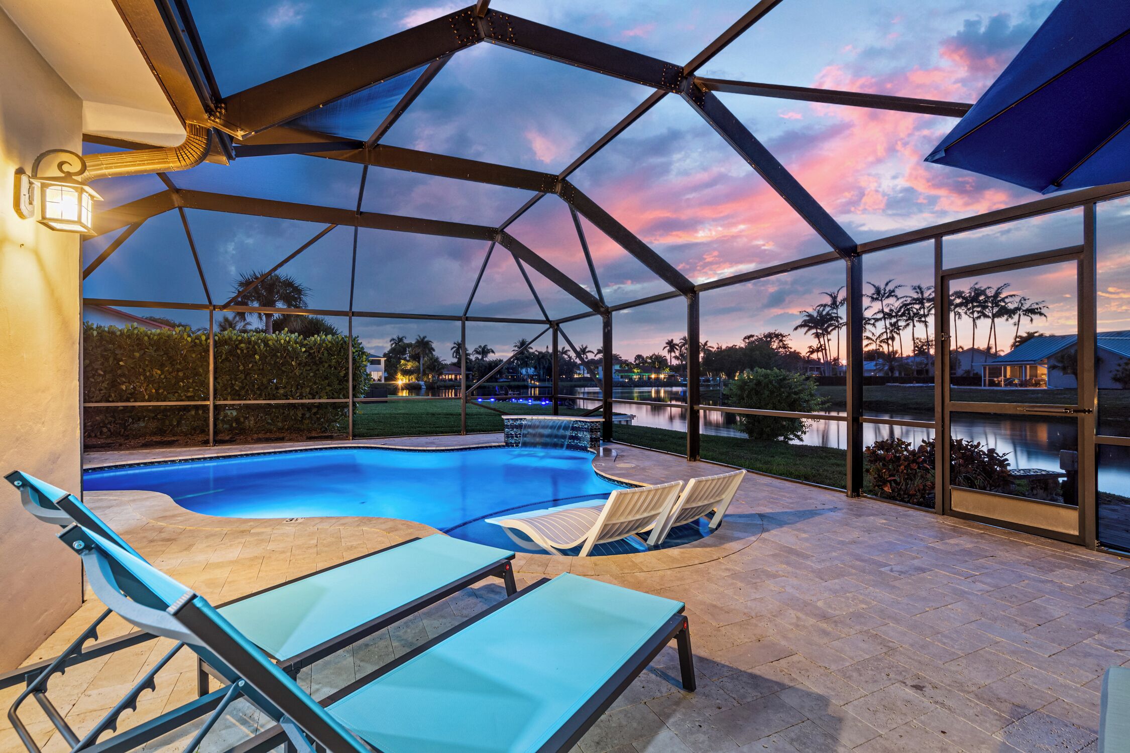 A covered outdoor swimming pool with lounge chairs and a view of palm trees and a lake at dusk.