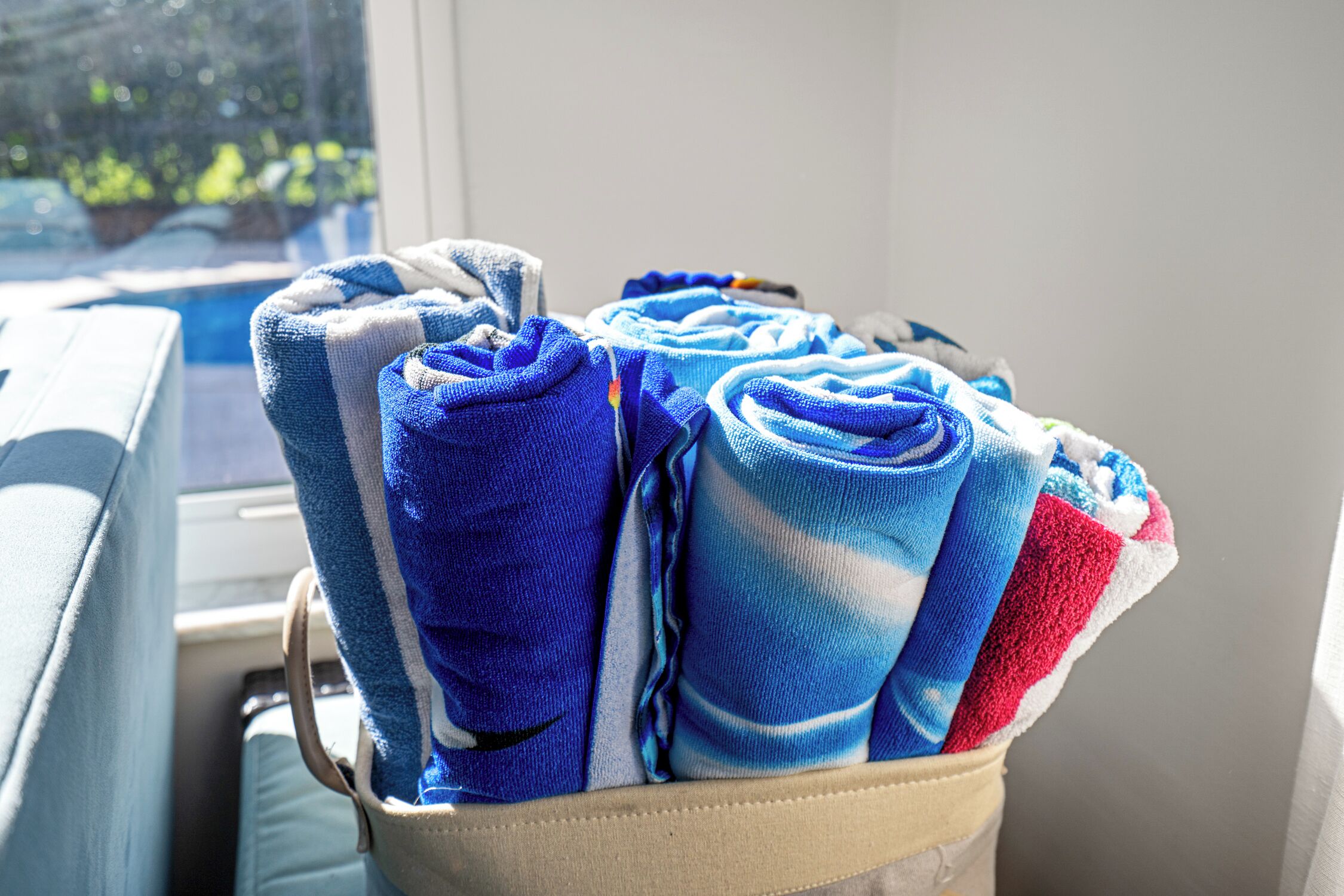 A window seat with a view of a pool and greenery, a window with blinds, and a bag of folded towels.