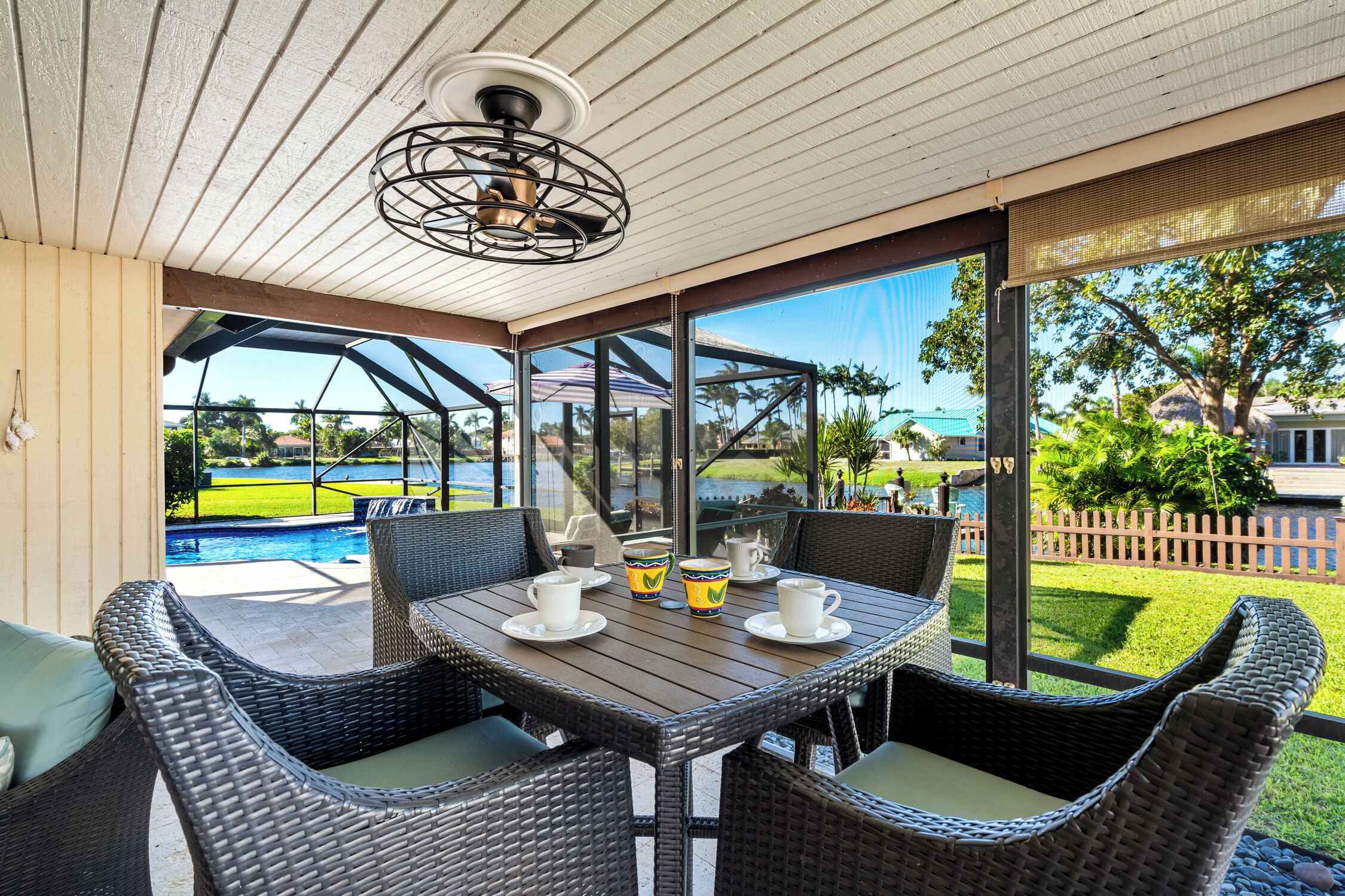 An outdoor patio with wicker furniture, a table with cups and a pitcher, and a view of a swimming pool and greenery.
