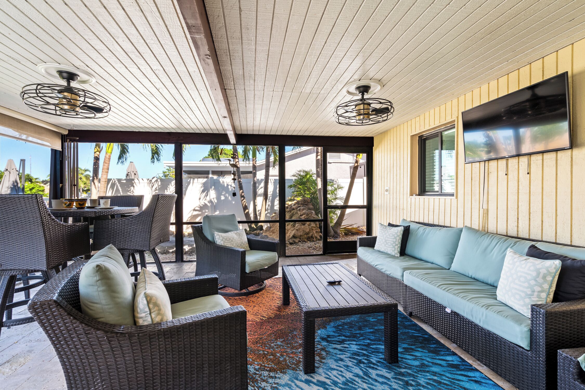 A covered outdoor seating area with wicker furniture, a flat-screen TV, and a view of palm trees and a building.