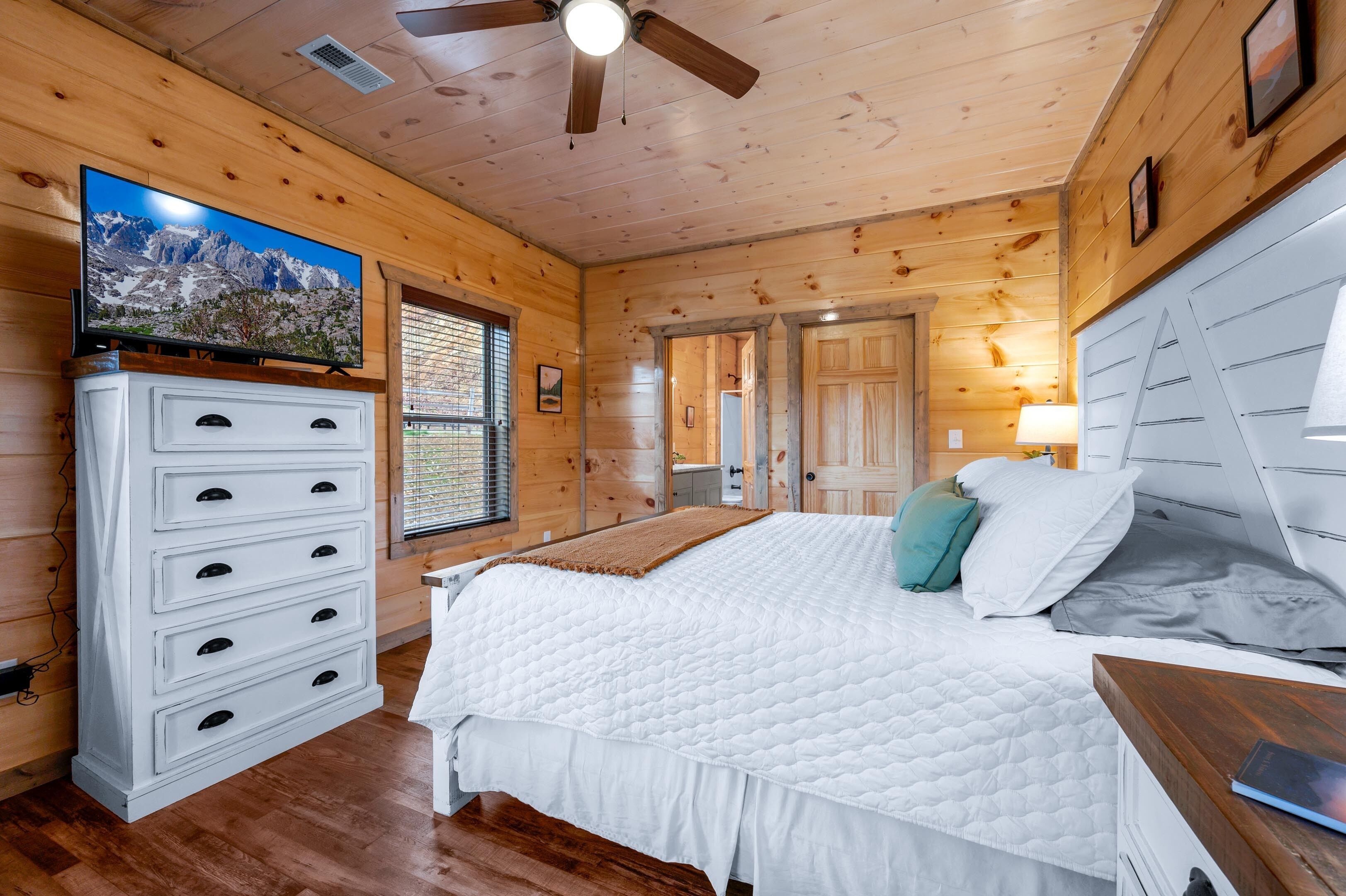 A wooden cabin bedroom with a bed, dresser, TV, and ceiling fan.