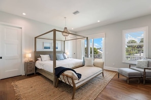 A four-poster bed with a canopy, a bedside table with a lamp, a chair, and a view of the outdoors through a window with blinds.