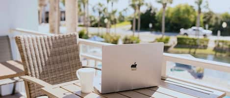 Ein Laptop steht auf einem Tisch neben einem Korbstuhl und einer weißen Tasse, mit Blick auf einen Pool und Palmen.