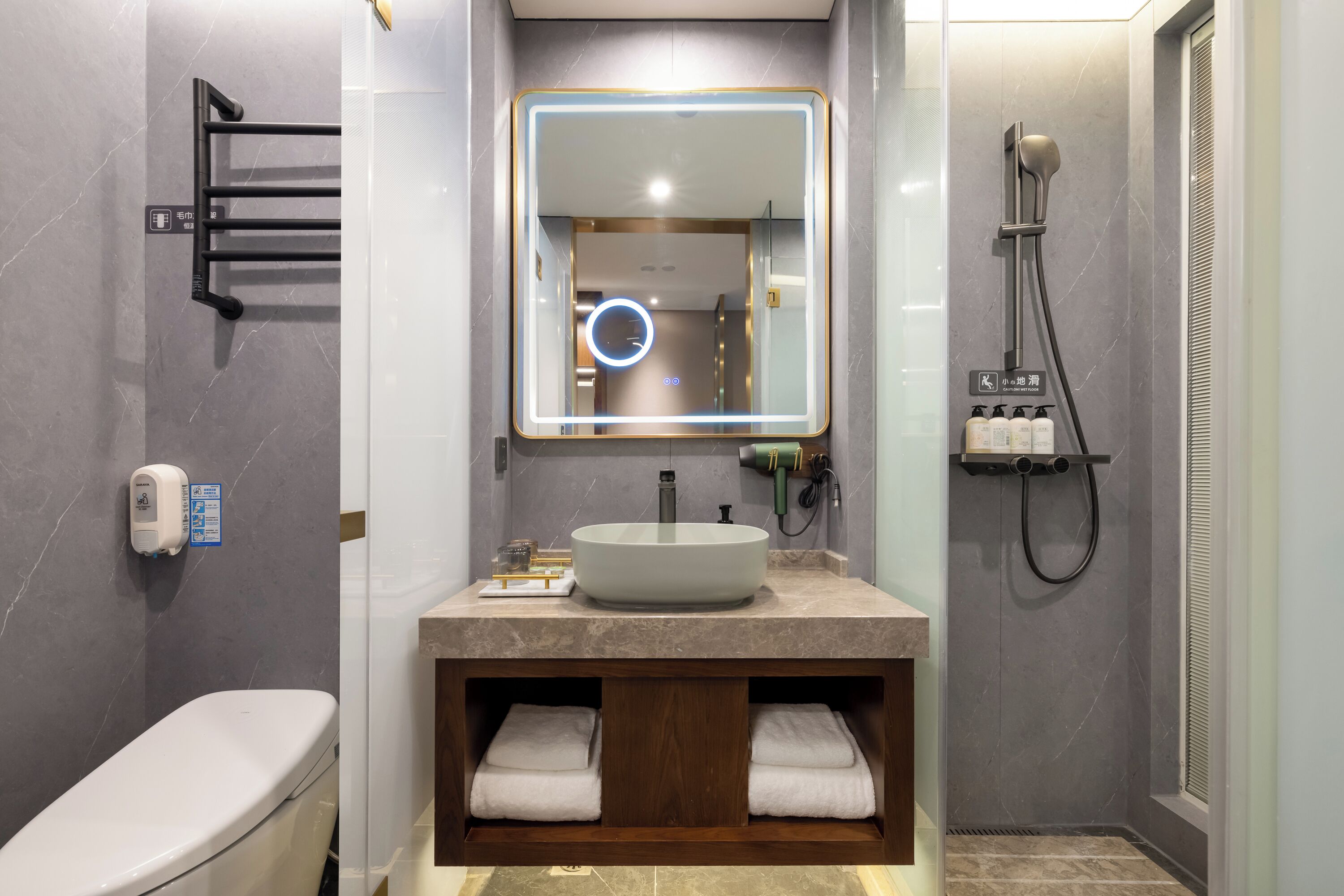 A modern bathroom with a toilet, a sink with a mirror, a towel rack, a handheld showerhead, and a soap dispenser.