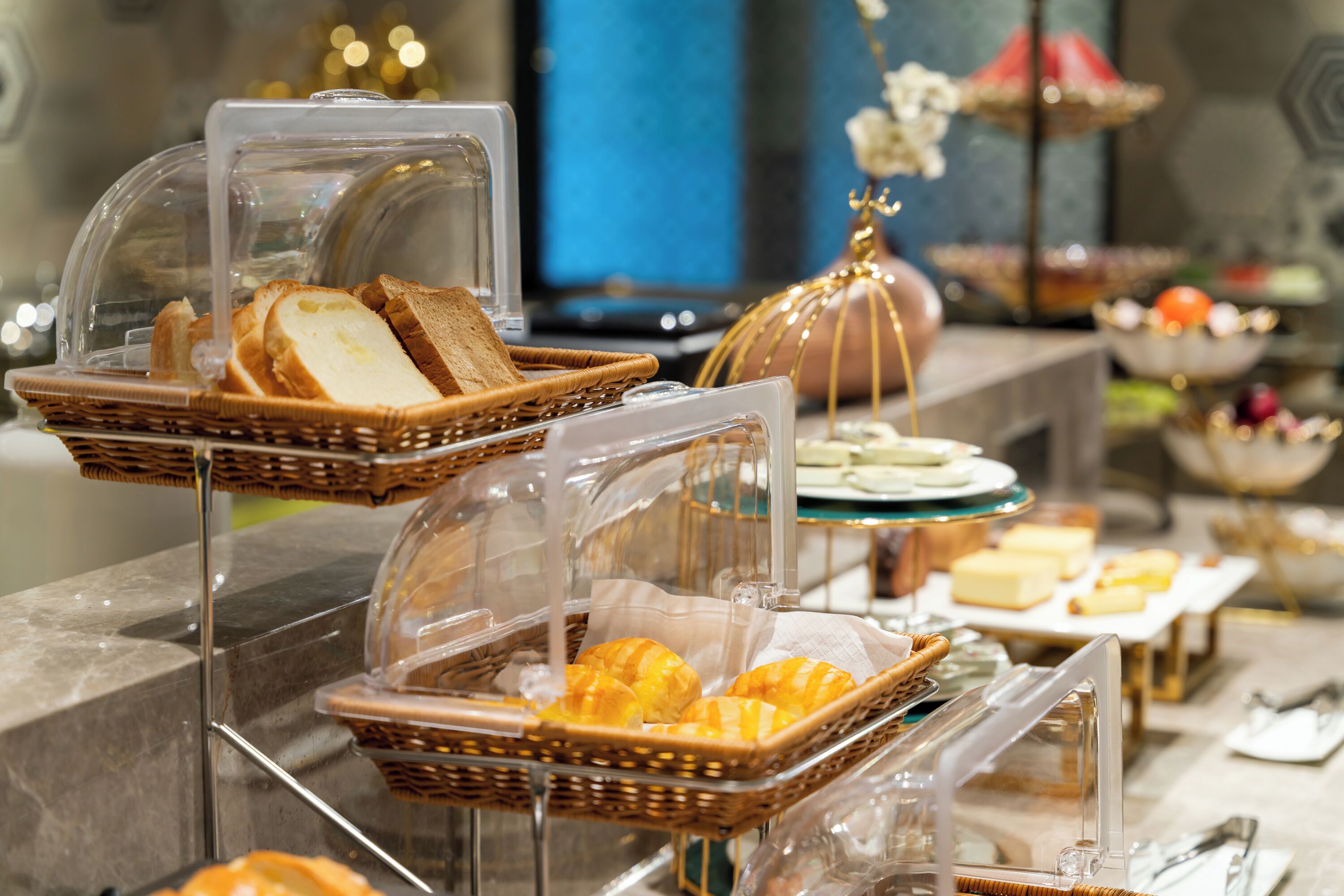 Tarjolla on buffet, jossa ruoka-annokset, kuten leipä ja leivonnaiset, sekä erilaiset juustot ja hedelmät on peitetty läpinäkyvillä muovikansilla.