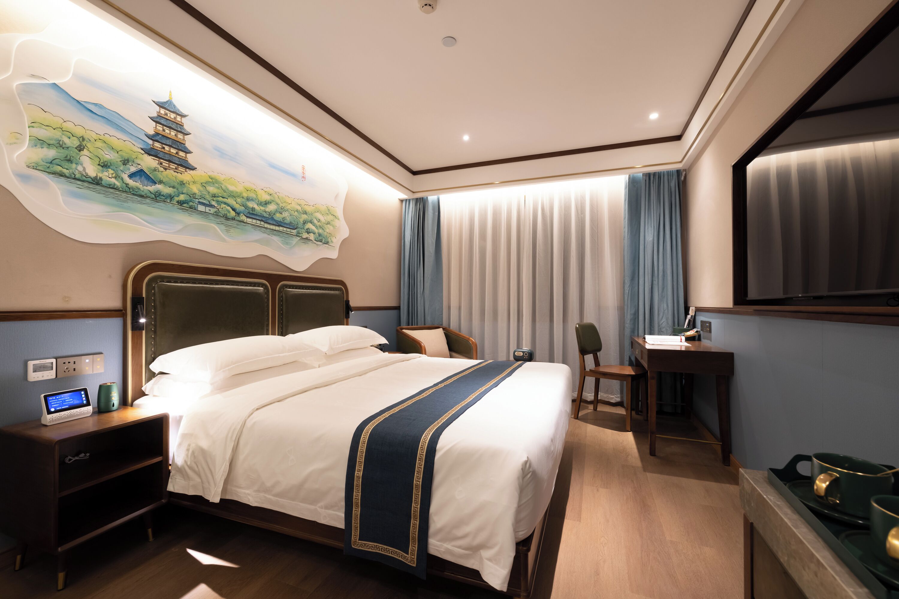 A hotel room with a bed, bedside table, nightstand, desk, chair, and a painting of a pagoda on the wall.