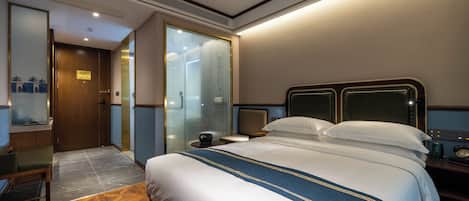 A hotel room with a large bed, a bedside table, a chair, a desk, and a bathroom with a glass door.