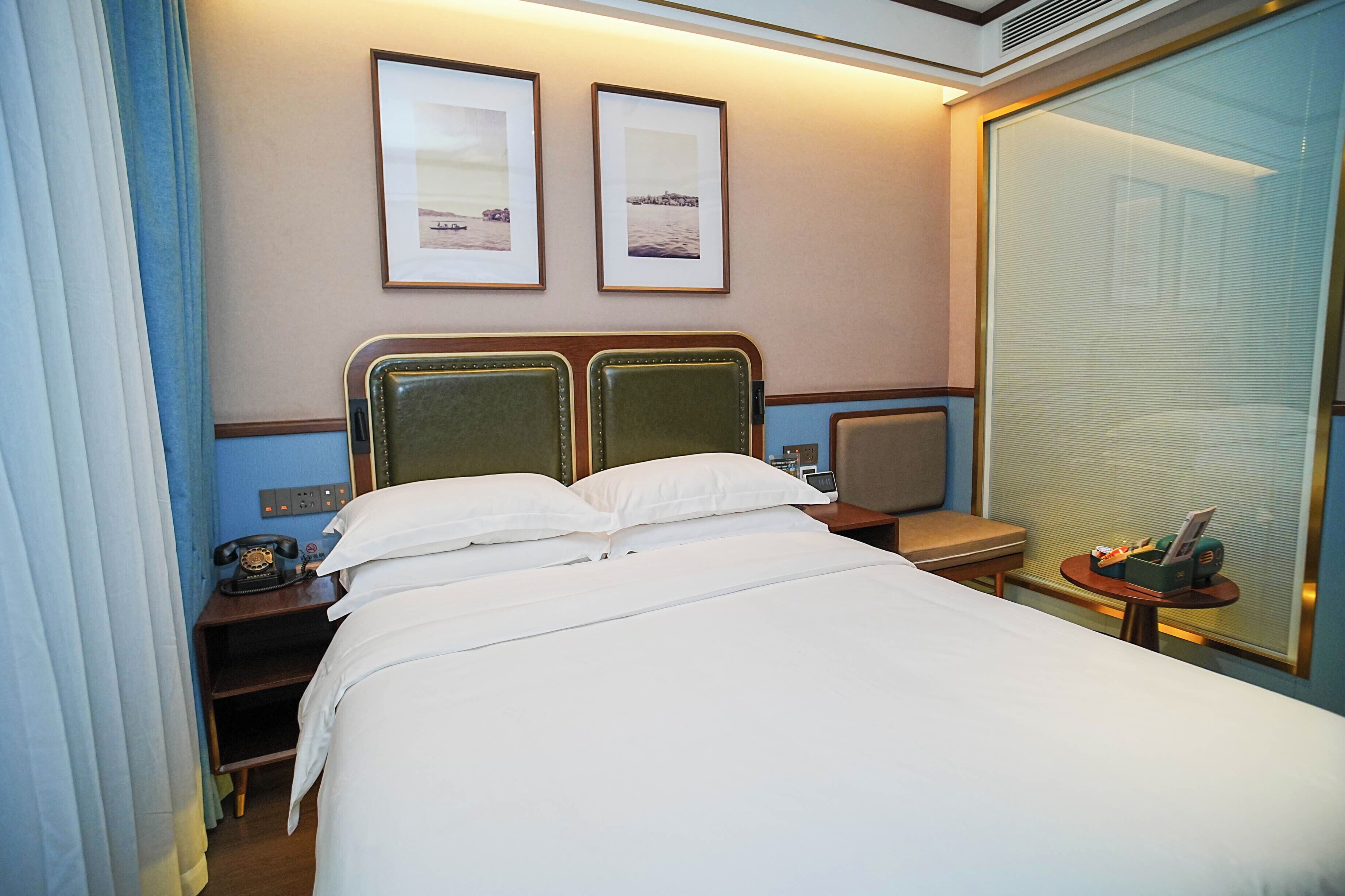 A hotel room with a large bed, two framed pictures on the wall, a bedside table with a telephone, and a small table with a magazine and a bottle.
