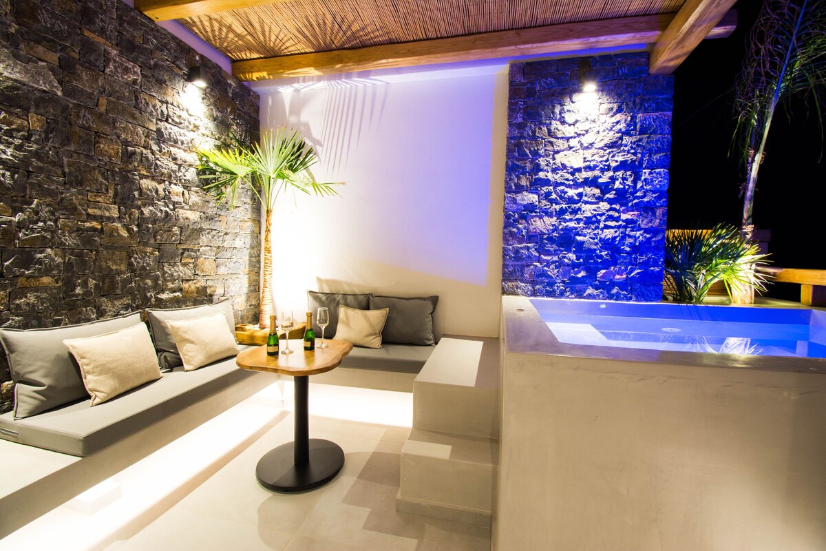 A modern lounge area with a stone wall, a sofa with cushions, a small round table with a bottle and glass, and a blue-lit water feature.