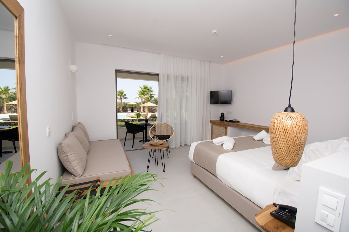 A modern hotel room with a bed, a desk, a chair, a TV, and a view of the outdoor pool area.