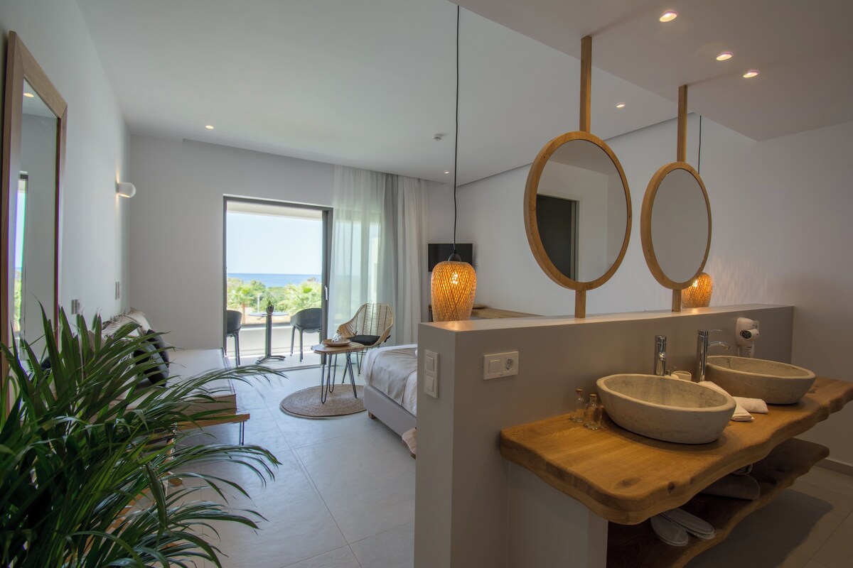 A modern hotel room with a large bed, a sitting area with a view, and a bathroom with two sinks and a large mirror.