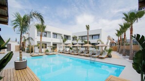 A swimming pool with lounge chairs and umbrellas, surrounded by palm trees and a modern building. - Aqua Blu (Hersonissos)