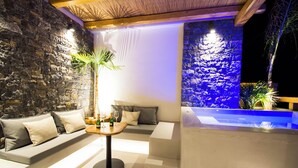 A modern lounge area with a stone wall, a sofa with cushions, a small round table with a bottle and glass, and a blue-lit water feature. - Aqua Blu (Hersonissos)