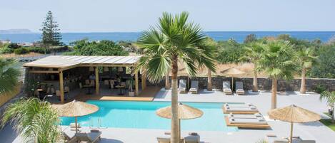 A pool area with lounge chairs and umbrellas, a bar area, and a view of the sea.