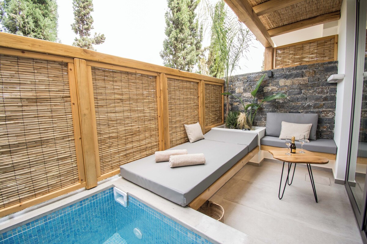 A modern outdoor relaxation area with a pool, a wooden deck, a stone wall, and a seating area with cushions and a table.