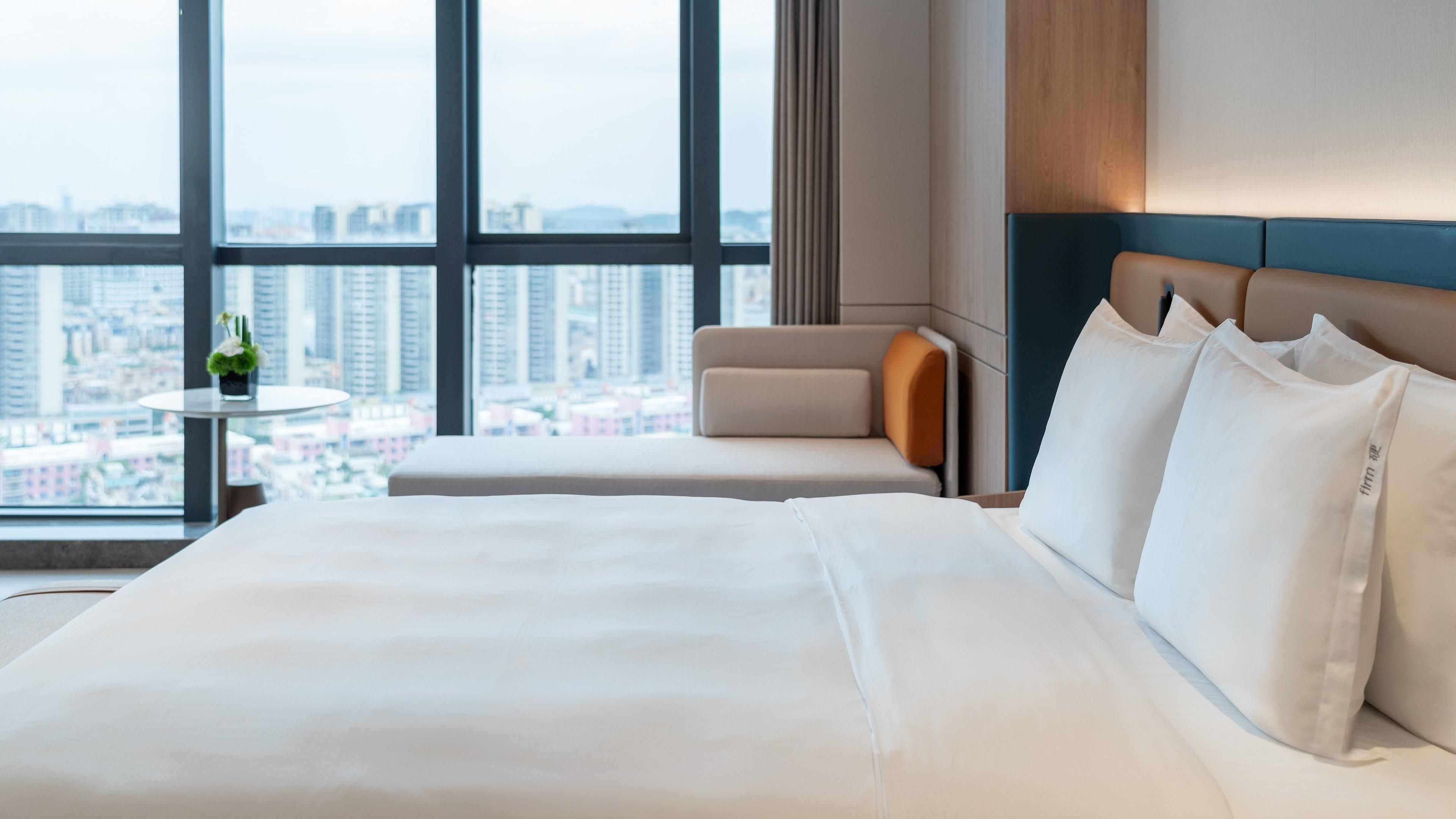A hotel room with a large bed, a small table with a plant, and a view of the city through large windows.
