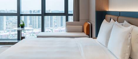 A hotel room with a large bed, a small table with a plant, and a view of the city through large windows.