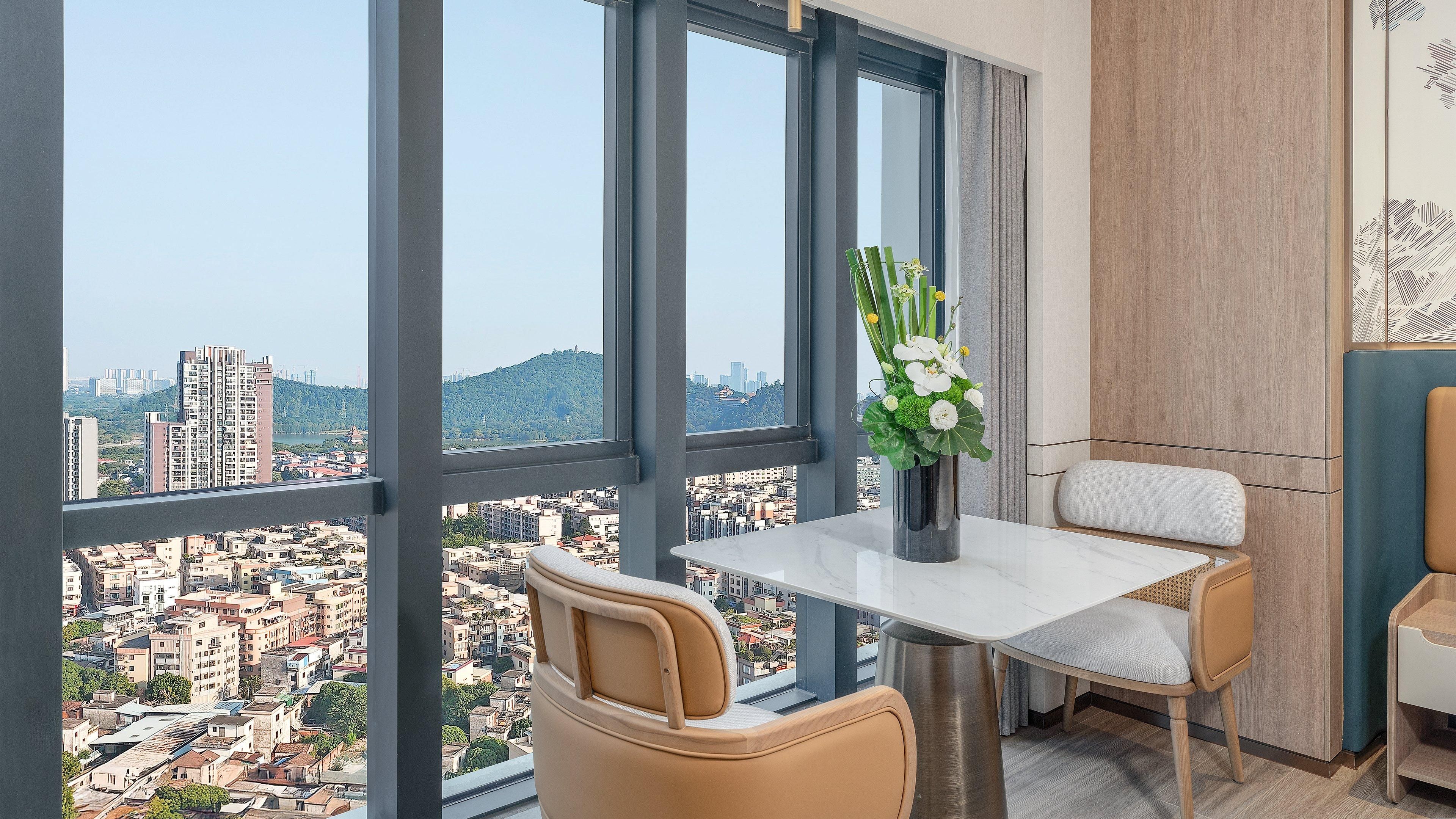 A modern room with a large window offering a city view, a small table with a vase of flowers, and a comfortable chair.