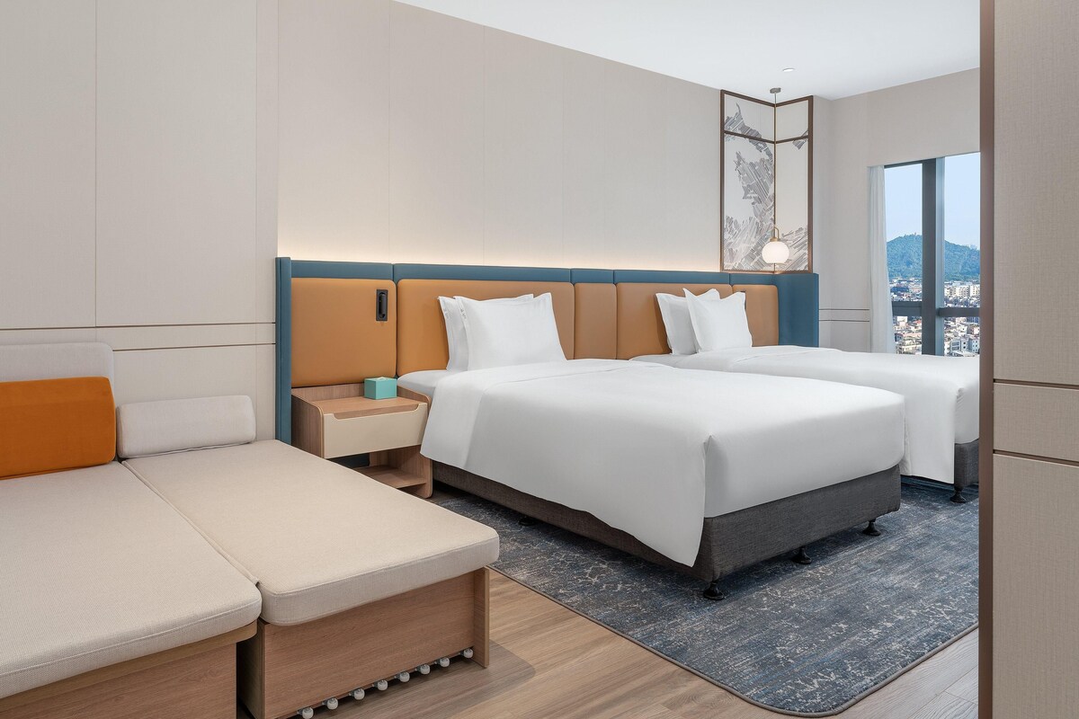 Ein Hotelzimmer mit Bett, Sofa, Nachttisch und Blick auf eine Skyline durch große Fenster.