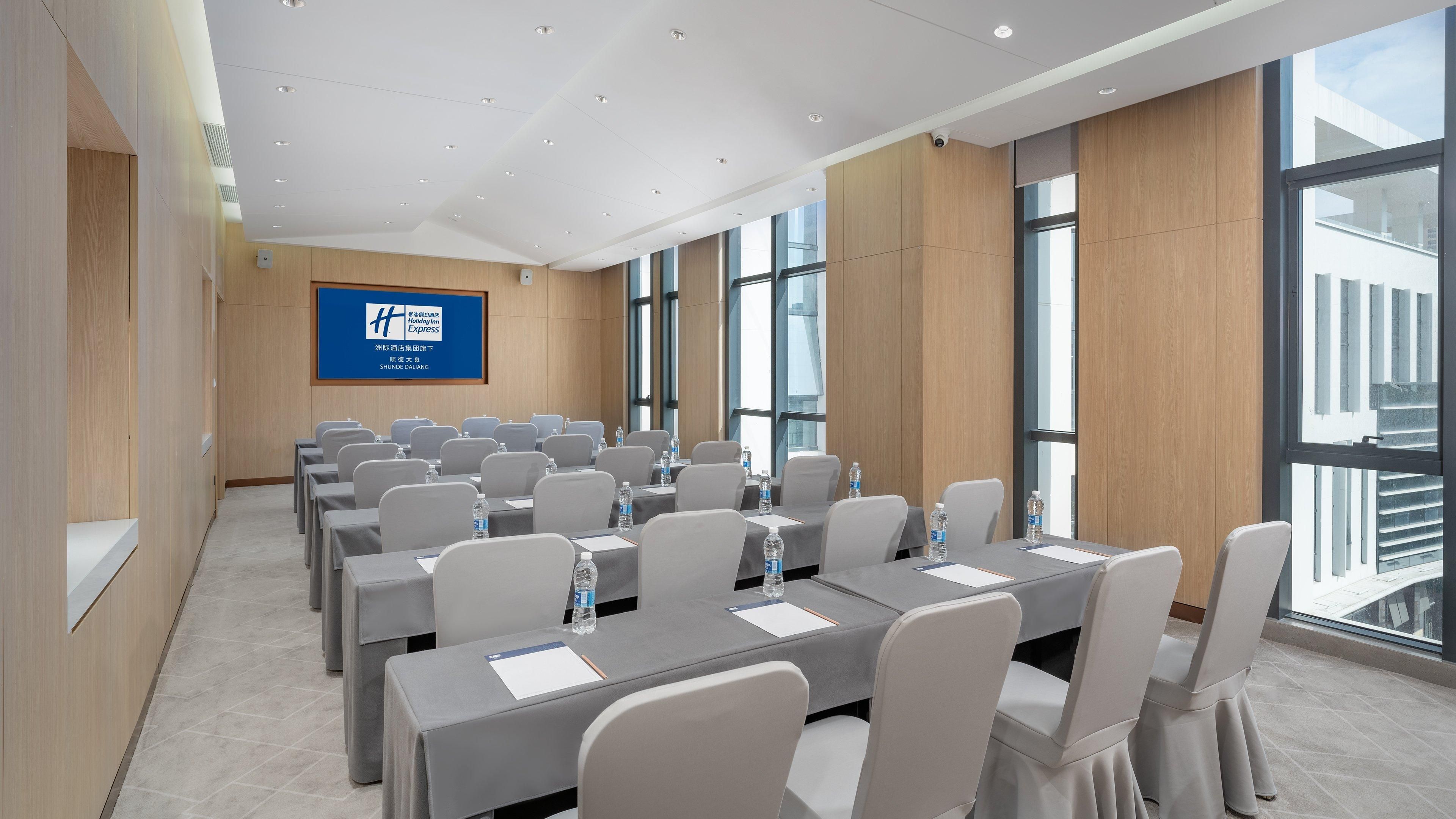 Un salón de conferencias con varias mesas y sillas, ventanas amplias y un letrero azul en la pared.