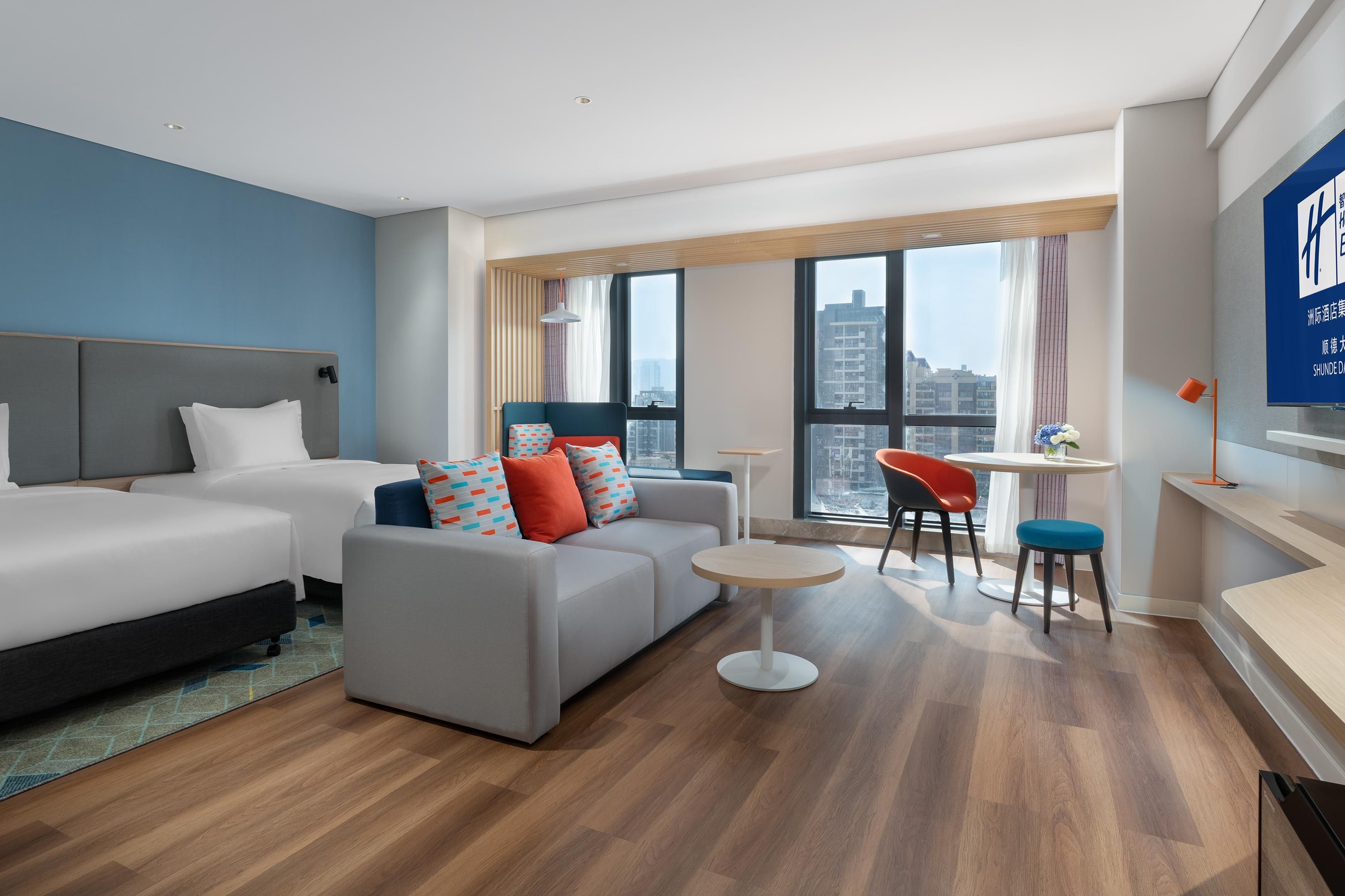 Een moderne hotelkamer met twee bedden, een bank, een klein rond tafeltje en uitzicht op de stad door grote ramen.