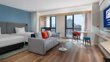 Een moderne hotelkamer met twee bedden, een bank, een klein rond tafeltje en uitzicht op de stad door grote ramen.