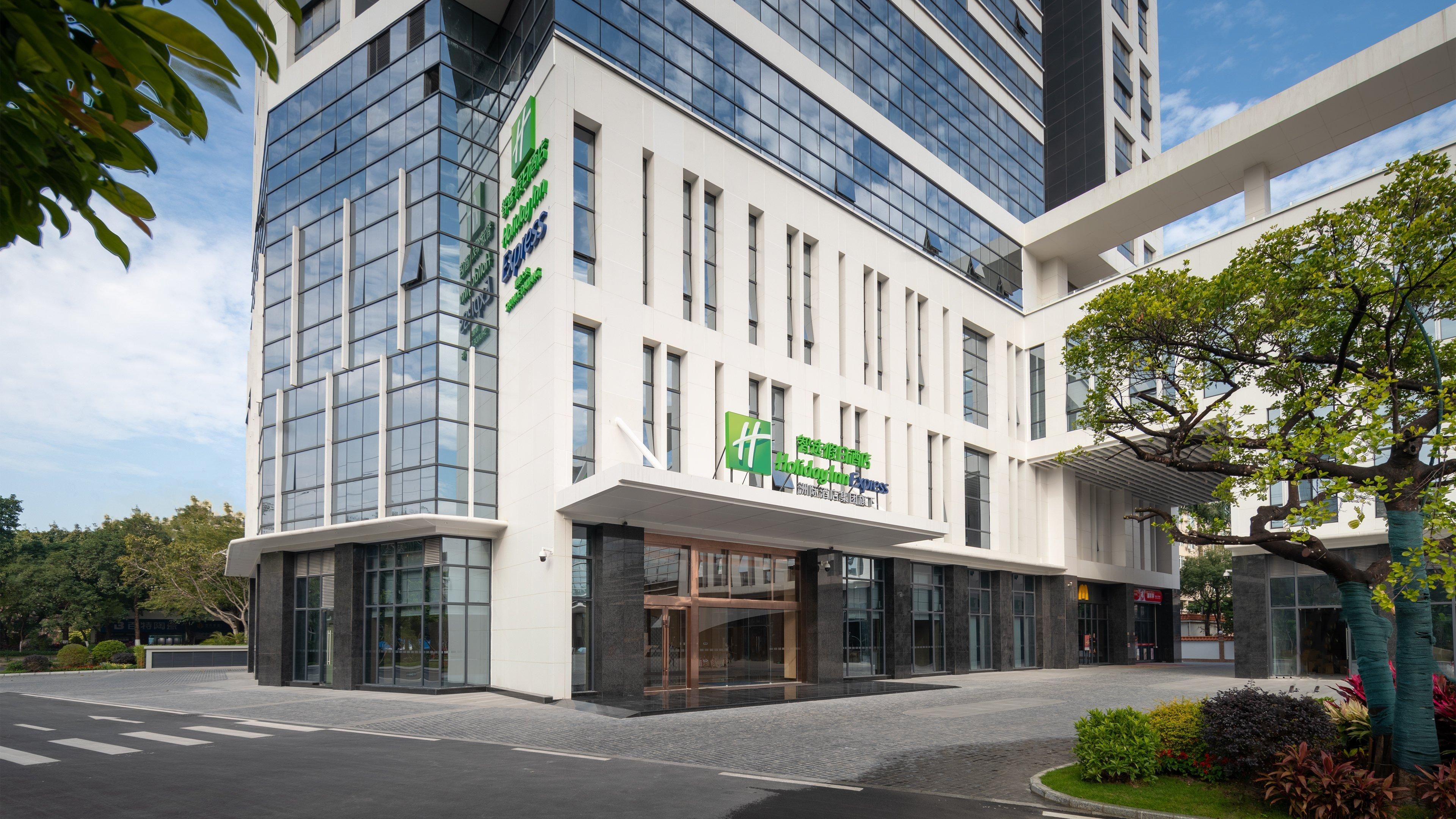 A modern hotel building with a glass facade, a prominent green and white sign, and a landscaped entrance area.