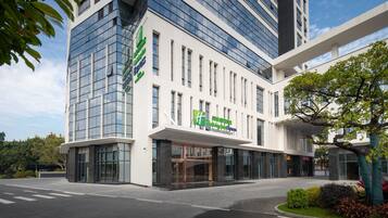 Un moderno edificio hotelero con fachada de vidrio, un llamativo letrero blanco y verde, y un área de entrada ajardinada.