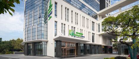 A modern hotel building with a glass facade, a prominent green and white sign, and a landscaped entrance area.