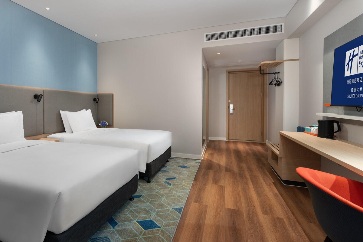 Ein Hotelzimmer mit zwei Betten, einem Schreibtisch, einem Sessel, einem Fernseher und einer Küchenzeile.