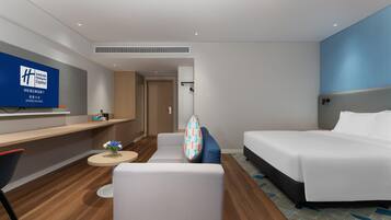 A hotel room with a bed, a sofa, a small table with a vase of flowers, a TV on the wall, and a wooden floor.
