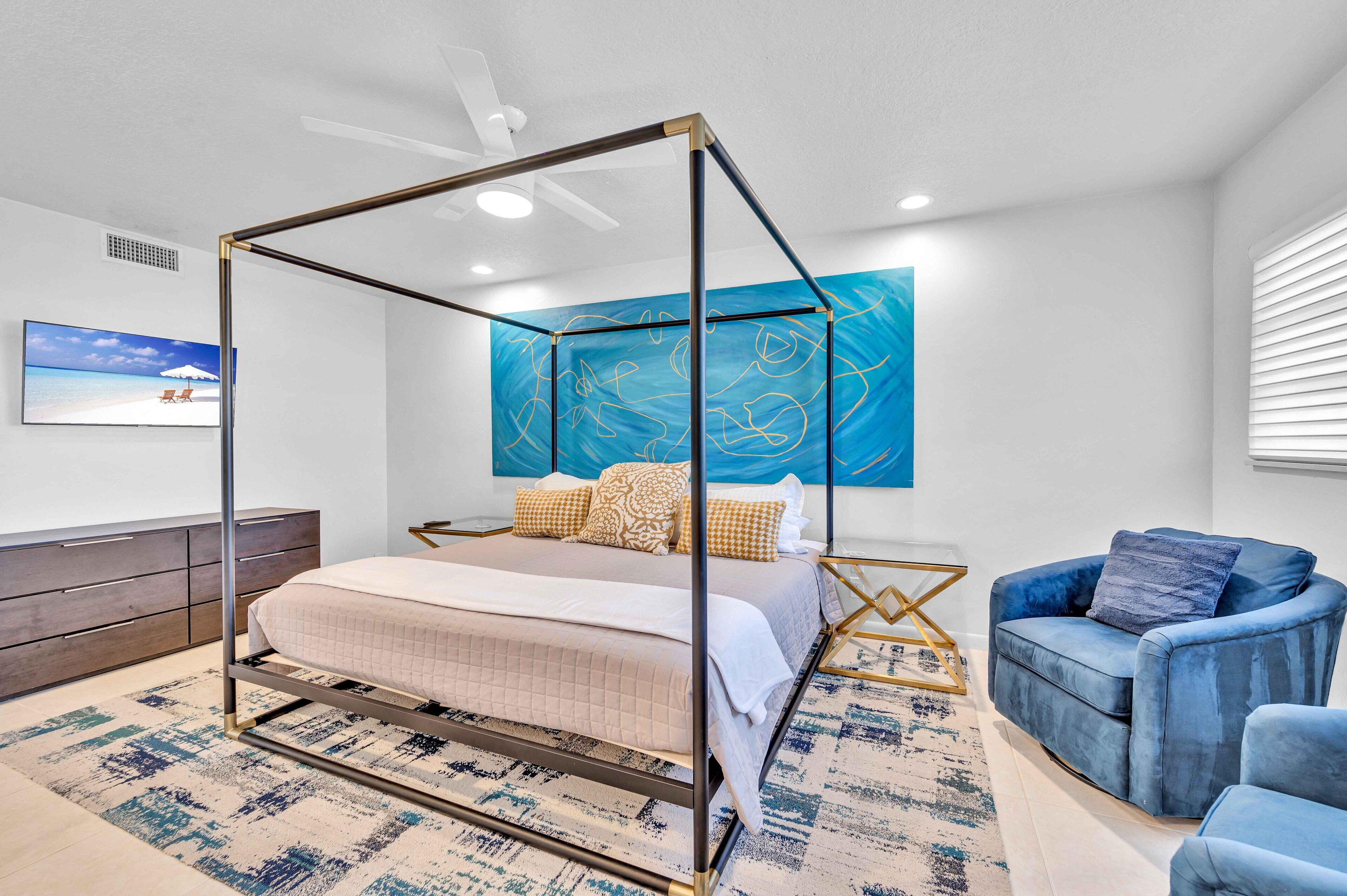 Ein Schlafzimmer mit Himmelbett, blauem Sessel, großem abstrakten Gemälde und Fernseher an der Wand.