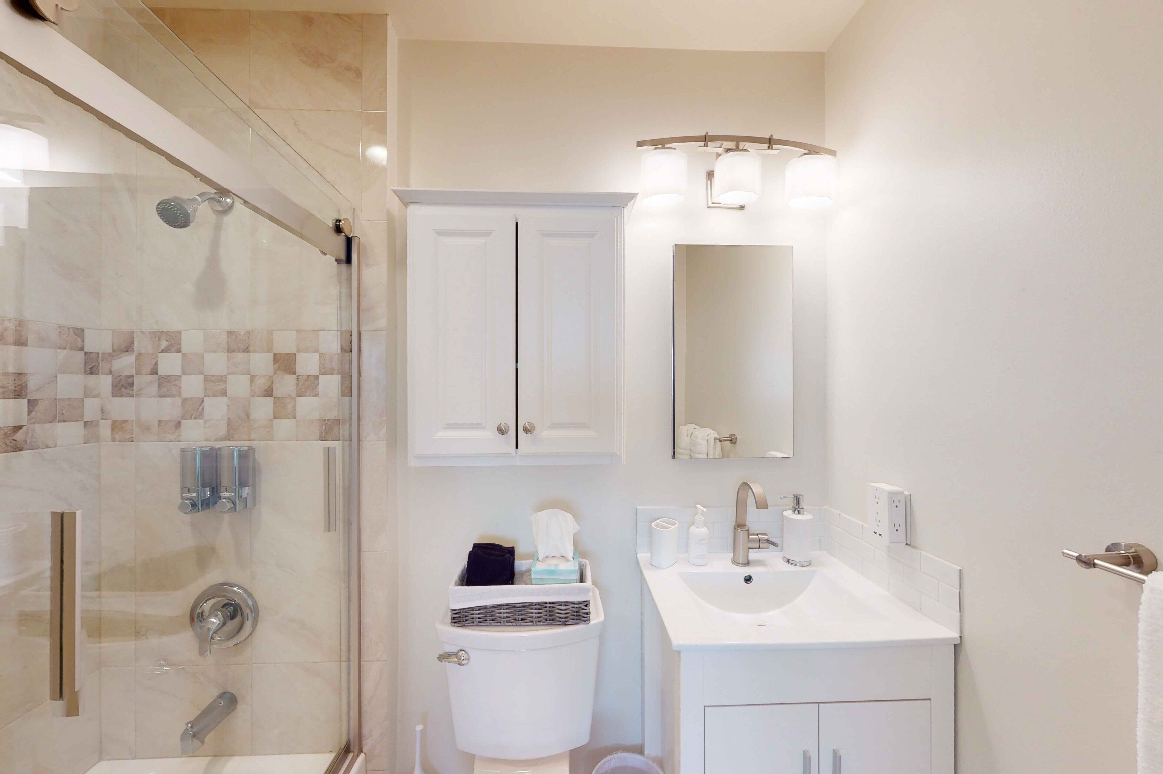A modern bathroom with a glass-enclosed shower, a white sink with a mirror above it, and a white toilet with a basket of toiletries.
