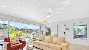 Ampla sala de estar com sofá bege, poltrona vermelha e ventilador de teto. Grandes janelas oferecem vista para uma piscina e vegetação.