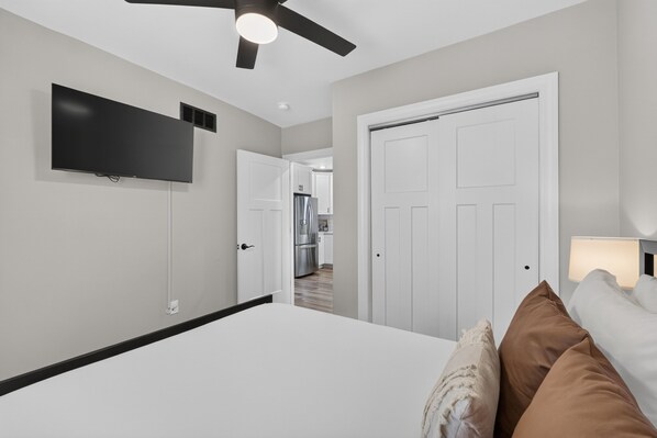 A hotel room with a bed, a flat-screen TV mounted on the wall, a ceiling fan, and a door leading to a kitchen area.