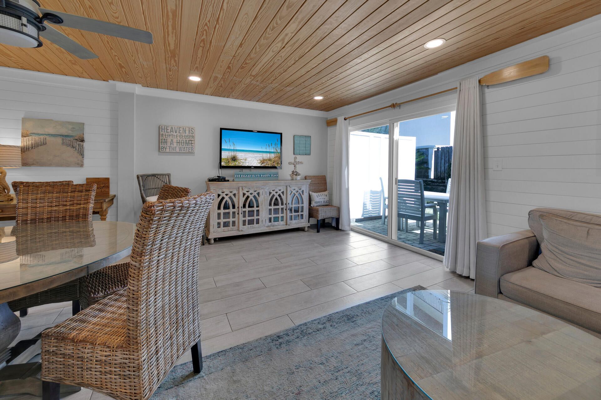 A spacious living area with a wooden ceiling, a glass coffee table, wicker furniture, and a view of the outdoors through a sliding glass door.
