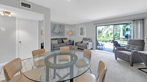 A modern living room with a glass table surrounded by chairs, a grey sofa set, a grey armchair, and a balcony with outdoor furniture.