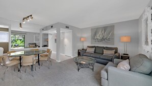 A modern living room with a glass dining table, grey sofas, and a large sailboat painting on the wall.