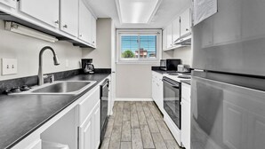 A modern kitchen with white cabinets, stainless steel appliances, a black countertop, and a window with a view of buildings outside. - Southbay by the Gulf 12 Overlooking Pool! ~ Short Walk to Beach! (Destin)