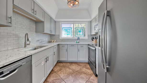 A modern kitchen with grey cabinets, a stainless steel refrigerator, and a window with a view of palm trees. - Southbay by the Gulf 124 | Beachfront Townhome w/ Pools & Patios (Destin)