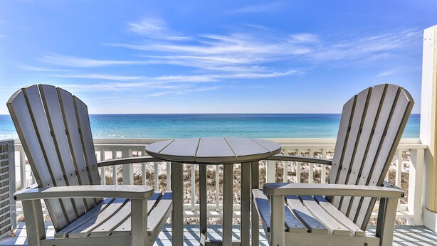 A deck with two Adirondack chairs and a small round table, overlooking a beach with clear blue water. - Completely Renovated 3-Bedroom Beach Front Townhome in Destin! (Destin)