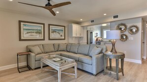A living room with a sectional sofa, a coffee table, a ceiling fan, and a kitchen in the background. - Completely Renovated 3-Bedroom Beach Front Townhome in Destin! (Destin)