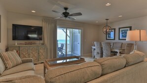 A living room with a flat-screen TV, a ceiling fan, wicker furniture, and a view to the outside.