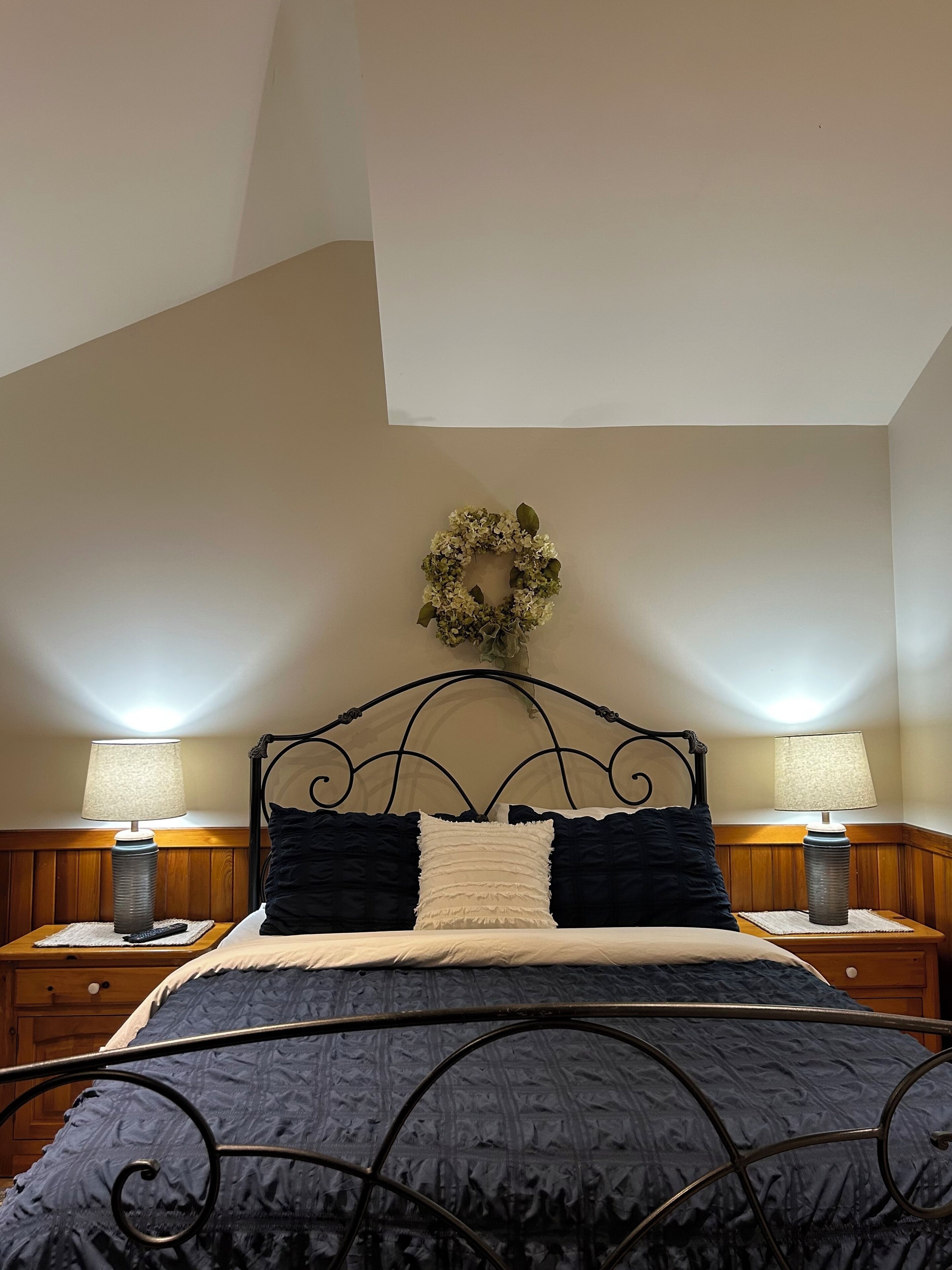 Een slaapkamer met een bed, twee nachtkastjes met lampen, een decoratieve krans aan de muur en een schuine plafond.