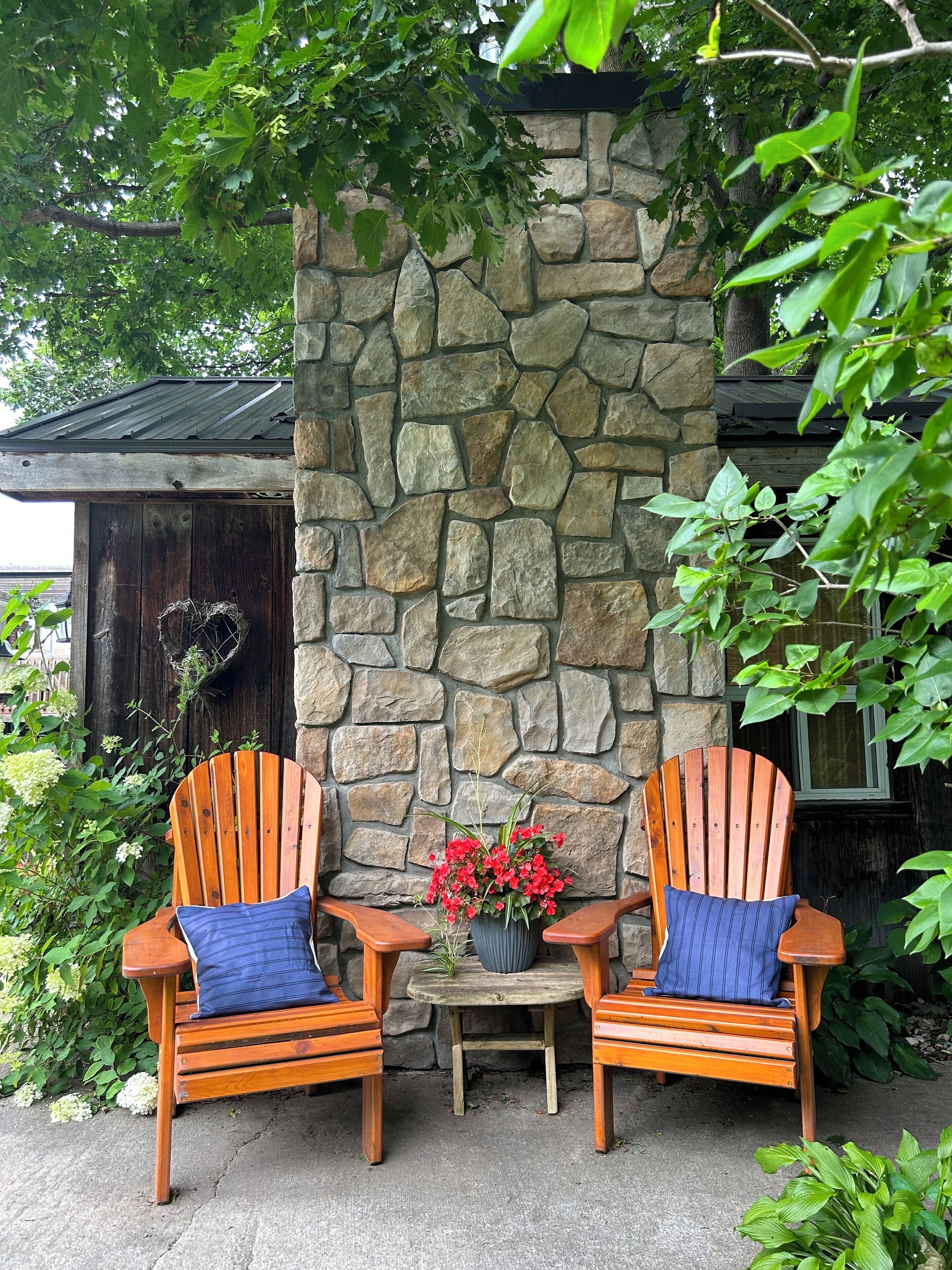 Two wooden Adirondack chairs with blue cushions, a stone wall, and a small table with a potted plant between them.
