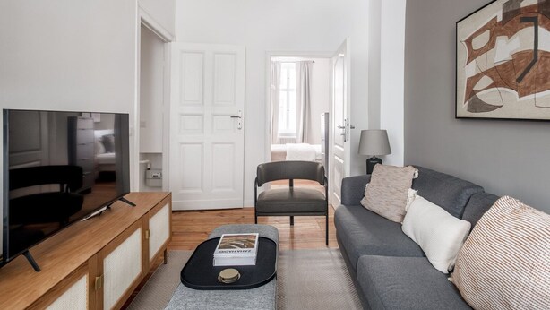 Una sala moderna con una televisión de pantalla plana, un sofá gris, una mesa de centro de madera y una puerta que da a un dormitorio.